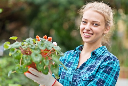 A young woman with blonde hair tied back, wearing a blue and green plaid shirt and gardening gloves, is standing outdoors in a lush garden. She is holding a small potted plant with green, rounded leaves and small white flowers, positioned in front of her chest. The background features a variety of greenery, including bushes and trees, with natural sunlight illuminating the scene. The garden appears well-maintained, with dense grass and a mixture of planted shrubs, suggesting a typical outdoor space that could benefit from professional gardening services offered by Landscapers Plaistow in the Plaistow area. The environment looks vibrant and healthy, reflecting care and attention to outdoor landscaping and garden maintenance, which are part of their expertise in gardening and lawn care.