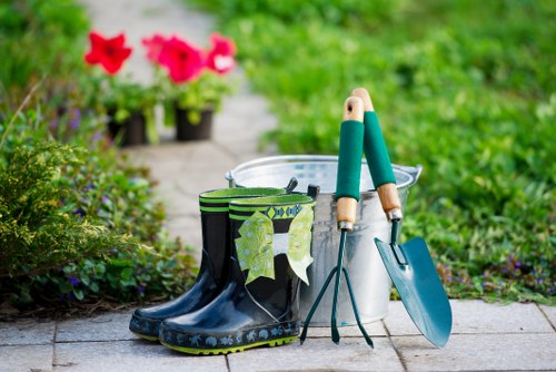 The image displays a small outdoor garden scene with gardening tools and accessories arranged on a paved pathway. In the foreground, a pair of black rubber gardening boots with a blue sole and decorative pattern sits next to a white plastic bucket, which contains a pair of green-handled shears. A garden trowel with a blue blade and a matching hand fork with wooden handles are also placed on top of the bucket, indicating preparation for garden maintenance. To the left, a green and black striped waterproof coat hangs over the boots. The background shows a lush, green garden with a variety of plants, including flowering pink geraniums in black plant pots, and a well-kept lawn area that extends into the distance. The scene appears to be set on a bright, mild day with natural daylight illuminating the space, emphasizing the vibrant greenery and natural textures of the garden surfaces. The setting suggests a typical landscaped yard with a mix of paved and planted garden elements, suitable for outdoor gardening activities or lawn care services offered by professionals such as Landscapers Plaistow.