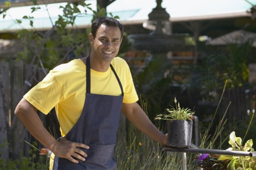 A male gardener with medium skin tone, wearing a yellow T-shirt and a dark blue apron, is standing outdoors in a landscaped garden in Plaistow. He is smiling and holding a black pot with a small green plant, positioned on a metal table or surface. The garden features a well-maintained lawn with lush, dense grass in the foreground, bordered by various shrubs and flowering plants. Behind him, there are trees and a wooden fence, with a garden shed or structure partially visible. The scene is illuminated by natural sunlight, suggesting a clear, bright day, with some shade cast by nearby trees. The overall setting indicates a professional gardening or landscaping environment, emphasizing outdoor care tasks that might be offered by Landscapers Plaistow, including plant maintenance and garden enhancement in a typical London suburb setting.