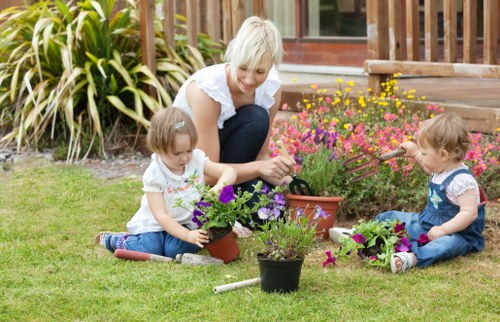 A mature woman with short blonde hair and wearing a white top is engaging in gardening activities with two young children on a well-maintained lawn. The children, both dressed in casual summer clothing, are seated on the grass, each holding small potted plants with colourful flowers, including purple and pink blooms. The woman appears to be helping or guiding the children in planting or potting the flowers. The garden features a lush green grass area with a tidy flower bed in the background, which includes a variety of flowering plants with pink, yellow, and green foliage, bordered by wooden decking. Large ornamental grass plants can be seen towards the back, partially obscuring a patio area with glass doors. The scene is depicted during daylight with bright, natural lighting, suggesting a clear and mild weather day, suitable for outdoor gardening. This setting showcases a tidy, landscaped garden space ideal for family gardening activities, and it aligns with gardening services such as lawn care, planting, and outdoor maintenance in the Plaistow area.