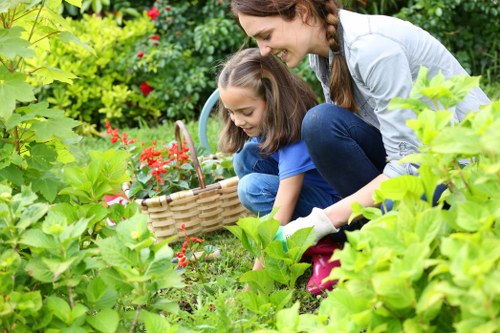 A woman and a young girl are engaged in gardening activities in a lush, well-maintained outdoor space in Plaistow, with a variety of green plants, shrubs, and flowering plants surrounding them. The woman is crouching down, smiling, and observing or assisting the girl, who is holding a small basket filled with red flowers or berries, possibly for planting or collecting. In the background, there are dense green hedges and trees providing shade, while the foreground features a neatly trimmed lawn with vibrant green grass. The scene is set on a bright, partly sunny day, emphasizing natural tones of green, red, and earth colours, and illustrating a friendly, productive outdoor environment indicative of professional gardening and landscaping services in the Plaistow area. The garden’s layout includes a mix of flower beds and grass patches, highlighting well-maintained outdoor spaces suitable for planting, lawn care, and outdoor family activities, paralleling the services offered by Landscapers Plaistow on their website.