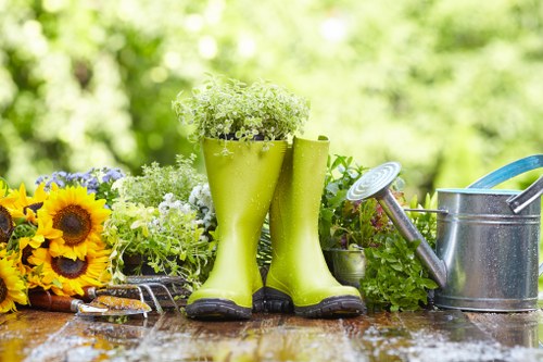 The image displays a garden scene with a weathered wooden outdoor table holding gardening tools and decorative elements. Prominently, there are a pair of bright green rubber rain boots filled with small white and green flowering plants, symbolising garden growth. To the left of the boots, a cluster of vibrant yellow sunflowers and other mixed flowering plants adds colour contrast, while on the right, a silver metal watering can with a blue handle and a spout is positioned, indicating active garden maintenance. The background features lush, out-of-focus greenery with trees and shrubs, suggesting a well-maintained garden space in a backyard or front garden setting. The scene is illuminated by natural daylight, with a soft, diffused light that highlights textures such as the smooth rubber of the boots, the metallic sheen of the watering can, and the glossy surface of the wet table, evoking a fresh, outdoor gardening environment typical of residential gardening services in Plaistow or nearby areas.