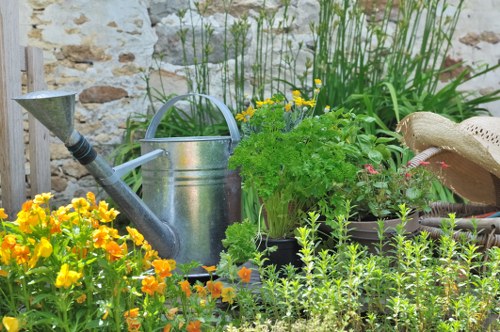 This image depicts a small, well-maintained garden scene featuring a variety of plants and garden elements. In the foreground, there is a lush patch of vibrant yellow and orange flowering plants with delicate blooms. Behind these, a taller, dense cluster of green foliage and herbs, including parsley or similar leafy herbs, is arranged in pots and planting containers. To the left, a traditional metal watering can with a rounded handle and spout is positioned among the plants, suggesting ongoing outdoor garden care. To the right, a straw hat rests on a surface, indicative of gardening activity. The backdrop includes a rustic stone wall, providing a textured, natural setting for the garden. The lighting suggests a bright, clear day, with soft natural light illuminating the greenery and flowers. This scene illustrates a typical garden space that may be part of a residential garden in Plaistow, showcasing seasonal planting and outdoor maintenance with a focus on plant health and aesthetic appeal, aligning with services offered by Landscapers Plaistow. The overall environment is peaceful, inviting, and suitable for standard gardening and landscaping activities.