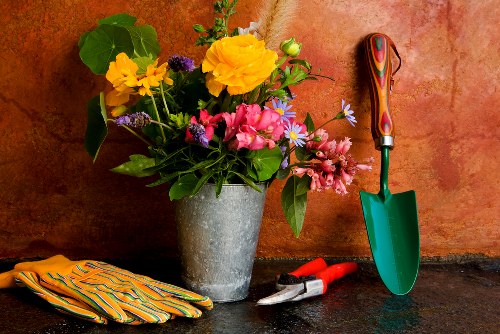 A close-up image of a gardening workspace featuring a large grey concrete planter filled with a colourful variety of fresh flowers, including yellow blooms, pink blossoms, and purple accents, set against a warm, textured terracotta wall. To the right of the planter, a small green trowel with a wooden handle is planted upright in the dark, slightly damp soil or work surface, with a pair of red pruning shears and a metal garden tool lying nearby. On the left, a pair of striped gardening gloves rest on the surface, indicating recent or ongoing gardening activity. The scene captures an outdoor or greenhouse environment with natural lighting, typical of garden maintenance and planting tasks, subtly aligned with professional gardening services offered by Landscapers Plaistow in the local area, such as Plaistow or surrounding parts of East London, ensuring visual relevance and natural connection to gardening and outdoor landscaping work.