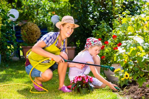 A woman and a young girl in a lush garden are engaged in gardening activities. The woman, dressed in a checkered shirt, yellow apron, and wearing a straw hat, is squatting and smiling as she holds a garden hose, facilitating watering. The girl, wearing a floral-patterned hat and light-colored clothing, is kneeling on the grass, carefully planting or tending to vibrant pink flowers in a flower bed bordered by a variety of colourful blooms, including red, white, and yellow flowers. The garden features a well-maintained grassy lawn, bordered by dense green foliage, shrubs, and small trees, with some decorative garden lights visible among the greenery. The scene is brightly lit, suggesting a clear, sunny day, and the environment exudes a warm, inviting outdoor space suitable for family gardening and landscape enhancement, consistent with services offered by Landscapers Plaistow servicing the London E13 postcode.