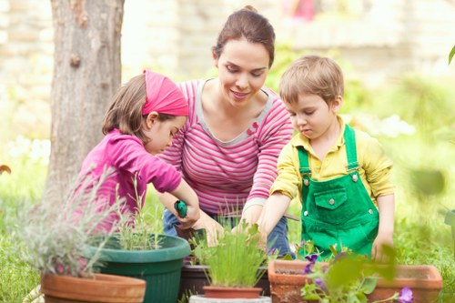 A woman with dark hair tied back, wearing a pink and white striped long-sleeve top, is gardening outdoors in a lush, green backyard with two young children. The girl on the left, wearing a pink headband and purple top, is watering plants in a turquoise pot, while the boy on the right, dressed in a yellow shirt and green overalls, is observing and assisting. The garden features a variety of potted plants, some flowering with bright purple and green foliage, arranged on a wooden surface. In the background, there are mature trees with textured bark and a blurred natural environment, indicating a well-maintained outdoor space in Plaistow. The scene is illuminated by natural daylight, suggesting a sunny day, and reflects activities typical of garden care and plant nurturing, demonstrating a family engagement focused on gardening, which aligns with landscaping and outdoor maintenance services offered by Landscapers Plaistow.