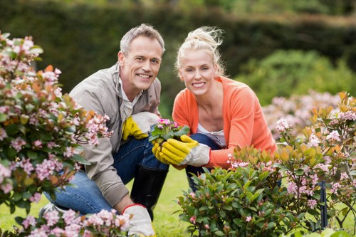 A man and a woman are kneeling together in a well-maintained garden, surrounded by lush green lawn, flowering shrubs with pink blossoms, and various other plants. The man has short grey hair and is wearing a beige jacket and yellow gardening gloves, holding a small potted plant. The woman has blonde hair tied in a bun, wearing an orange long-sleeve top and also sporting gardening gloves, smiling as they work together. The garden features a neatly trimmed hedge in the background, with a backdrop of trees and a slightly overcast sky, suggesting mild weather. The scene captures a moment of outdoor gardening activity, emphasizing careful plant inspection and care, which aligns with the gardening services offered by Landscapers Plaistow in the Plaistow area, located near E13 postal code. This natural, vibrant setting highlights the importance of garden maintenance and horticultural care in local outdoor spaces.